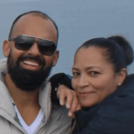 The image shows two people posing for a photograph with a backdrop of a body of water.