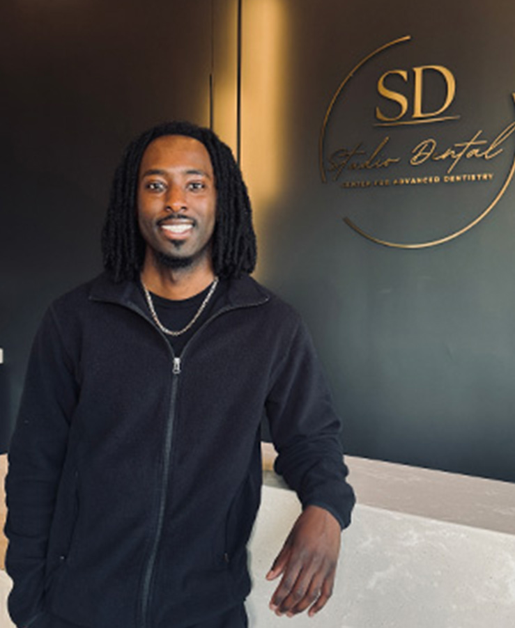 A man poses in front of a sign with the initials SD on it, standing against a wall with a logo and his hands in his pockets.