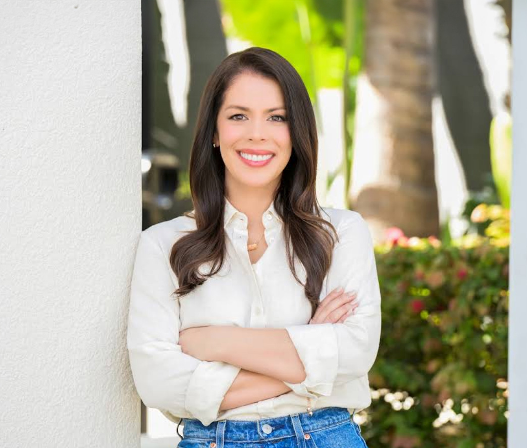 The image features a woman posing with her arms crossed, standing against a wall with plants behind her, smiling at the camera.