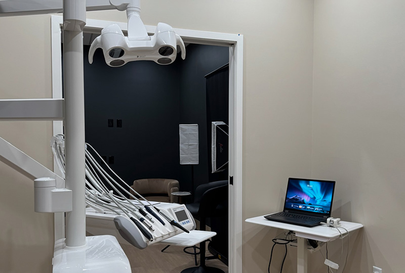 The image shows a dental office interior with a modern dental chair, a computer screen displaying medical images, and a desk with a laptop and monitor.