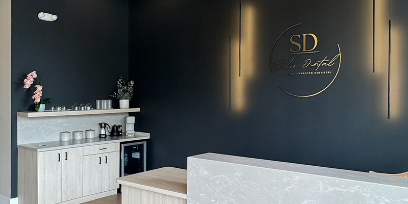 The image shows an interior space with a dark background featuring a modern design, including a countertop with a logo, a lighted display case, and decorative elements such as framed pictures on the wall and a potted plant.