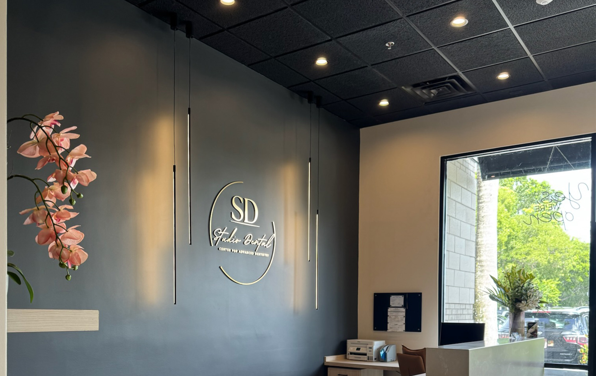This is a photograph of an interior space featuring a modern decor style with a black wall, illuminated signage, framed artwork, and a floral arrangement on a table. The room has a clean, minimalist aesthetic with a focus on neutral colors and contemporary design elements.