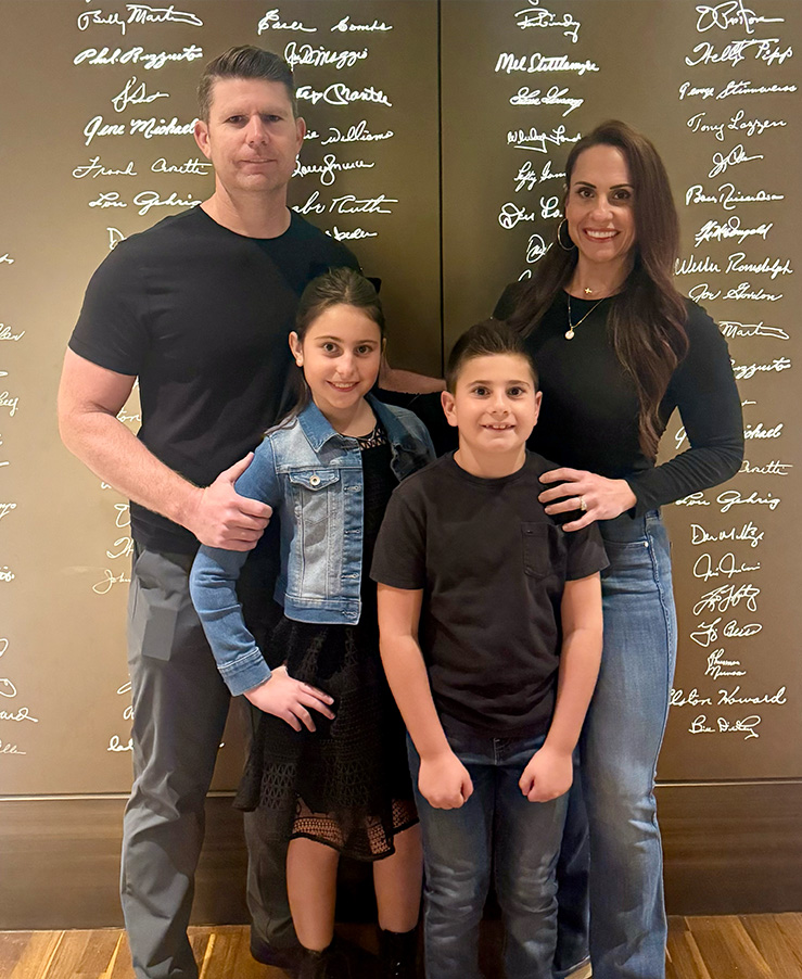 A family posing together with a man, woman, and two children in front of a wall displaying autographs.