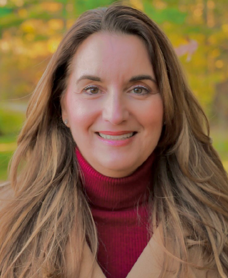 A woman with long hair wearing a red sweater over a white turtleneck, standing outdoors against an autumnal background.