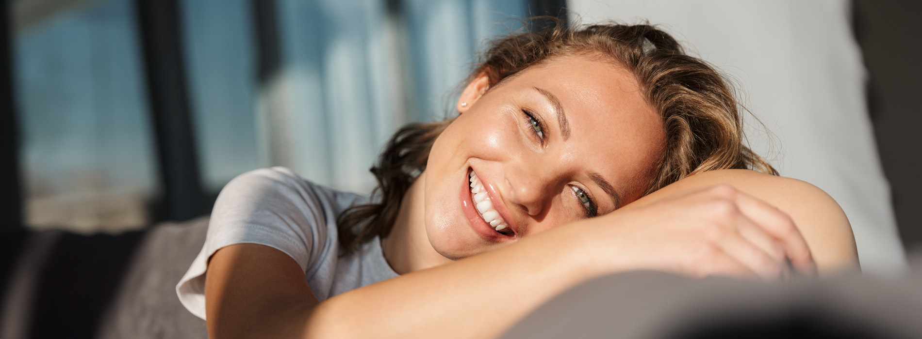 A woman with a radiant smile is seen resting her head on her hand while lying down on a bench outdoors, enjoying a sunny day.