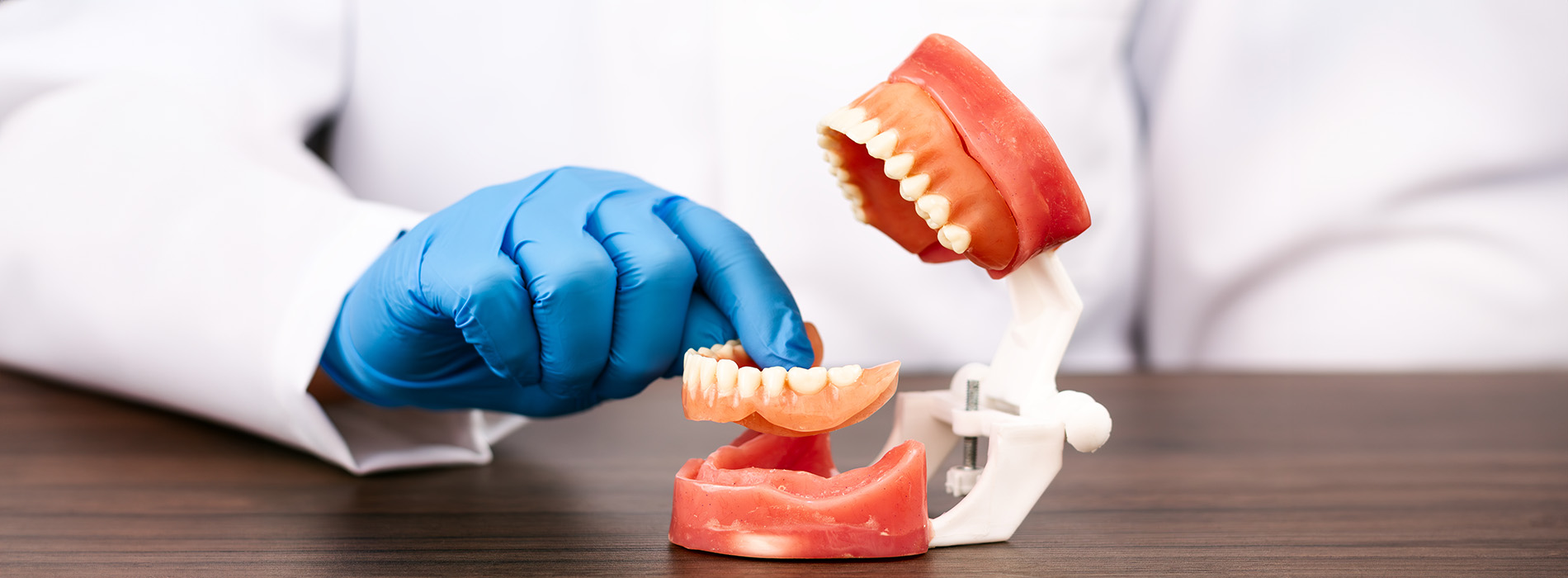 The image shows a table with a toothbrush and a small blue robot figurine positioned next to each other, with the robot figurine holding a toothbrush, set against a blurred background that appears to be a room with a white wall and some indistinct objects.