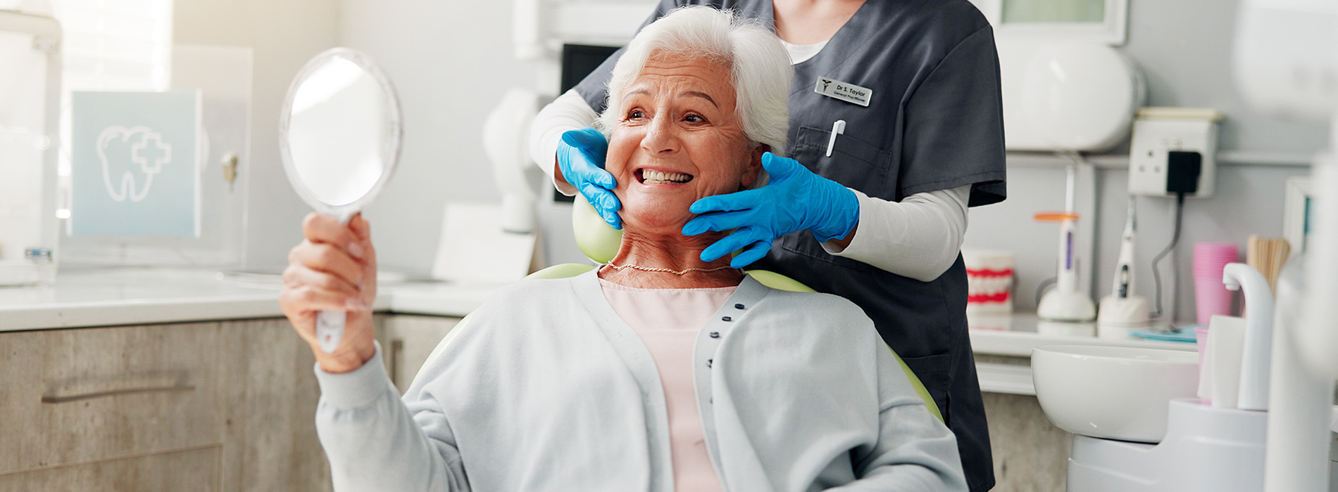 An elderly woman receiving dental care from a professional in a modern dental office, with a smiling expression and holding up her hand in a gesture of approval.