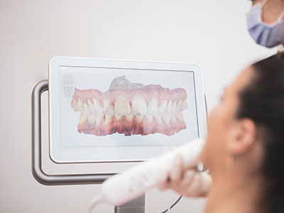A photograph showing a person using a digital dental imaging system with a 3D model of a mouth displayed on its screen, likely for dental treatment planning or oral health analysis purposes.