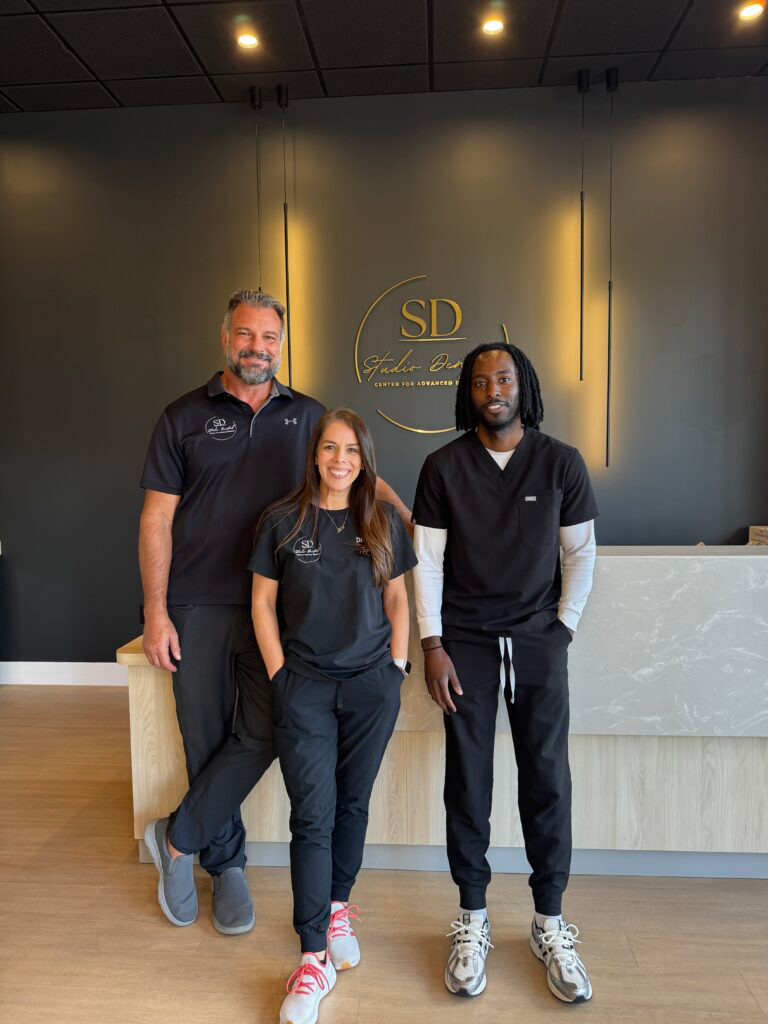 In the image, there are three individuals posing in front of a sign with the initials SD on it, which stands for Sports Dentistry . The person on the left is wearing a uniform with the logo of a sports team, the middle individual appears to be a dentist, and the person on the right is dressed in casual attire. They are all standing in front of a desk with a marble top and displaying their smiles for the photograph.