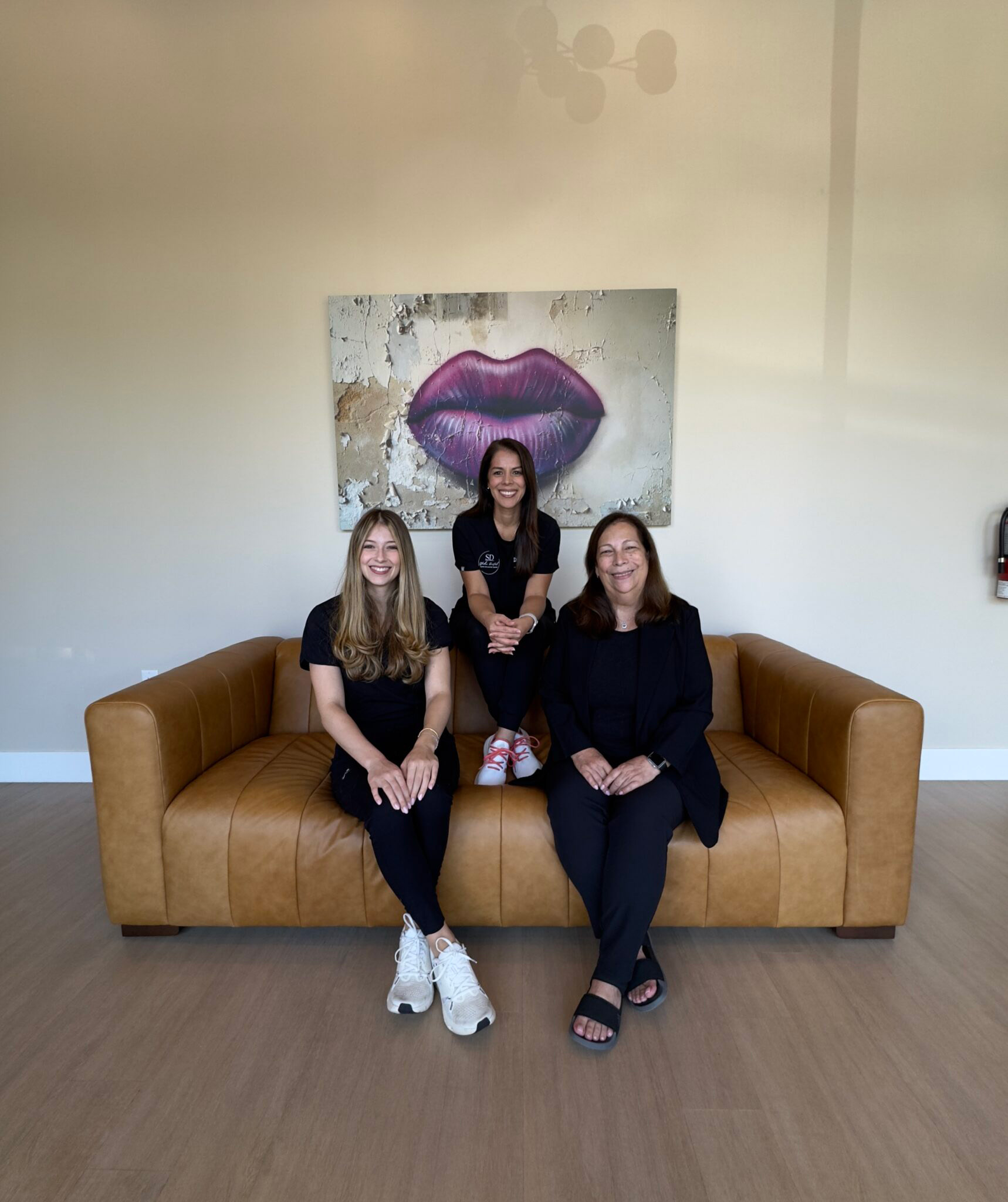 Three individuals posing together in front of a painting on a wall, with two women seated on a couch and a man standing behind them.