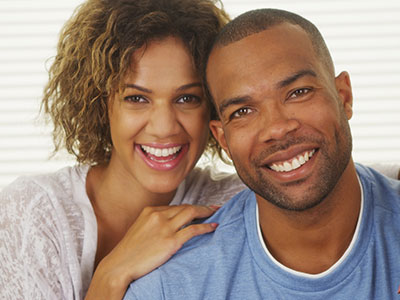 This is a color photograph featuring two adults, a man and a woman, smiling at the camera. The man has short hair and appears to be wearing a casual blue shirt, while the woman has curly hair and is wearing a white top with lace sleeves. They are standing close together with their arms around each other, suggesting a close relationship.