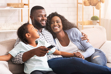 The image shows a family sitting together on a couch, with the father holding a remote control, smiling at the camera, while the mother and child look towards him. They are all looking happy and relaxed.