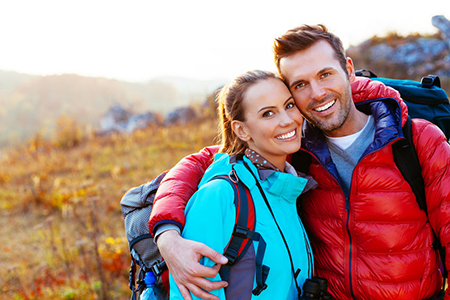 A couple posing for a photo outdoors with backpacks, smiling at each other.