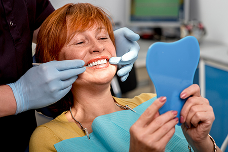A woman sitting in a dental chair with a blue toothbrush-shaped mouthguard over her teeth, smiling, while holding up a mirror with a similar mouthguard to show it off, with a dental hygienist assisting behind her.