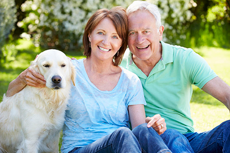 This is a color photograph featuring an older couple sitting outdoors with a golden retriever dog between them. The woman has short hair, wears a blue top, and smiles at the camera  the man has graying hair, is wearing a green shirt, and also smiles. They appear to be enjoying a pleasant moment together.