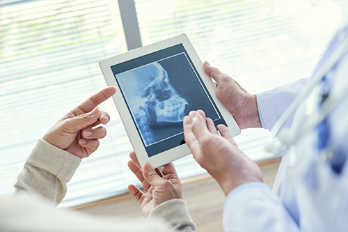The image shows a person holding a tablet displaying an X-ray, with another individual pointing at the screen while standing beside them, both are in a medical setting, possibly a hospital or clinic.