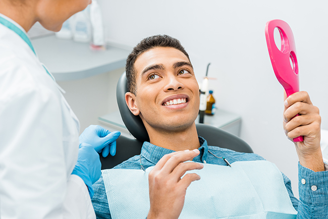 The image shows a man sitting in an dental chair with a smiling expression, holding a pink object that resembles a toothbrush, while a dentist attends to him.