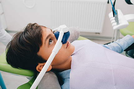 The image shows a person sitting in a dental chair with a dental mirror on their face, receiving dental care in an indoor setting.