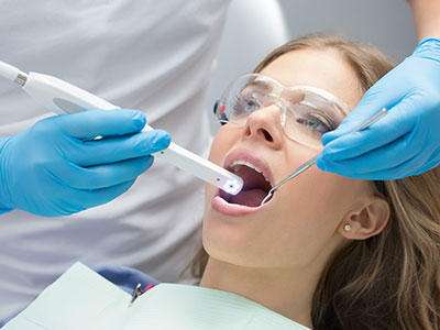 A woman receiving dental treatment with a dental device inserted into her mouth, attended by dental professionals wearing blue gloves and using a digital device to monitor the process.
